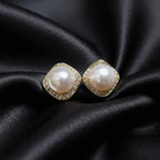 Pair of gold-tone cushion-square halo studs with round white centers, resting on black folded fabric.