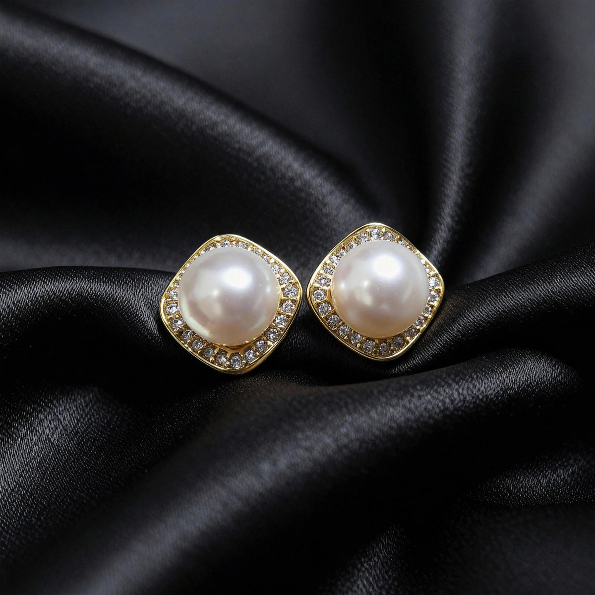 Pair of gold-tone cushion-square halo studs with round white centers, resting on black folded fabric.