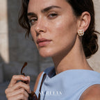 Close face-and-ear portrait holding sunglasses; white four-lobed stud, central bead and stone-lined inner outline are clear.