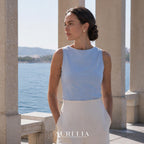 Standing waterfront portrait between pale stone columns, wearing the white and gold-tone clover stud.