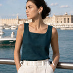 Waist-up model in a teal top and pale trousers beside a marina, with the bar and gray bead earring visible.