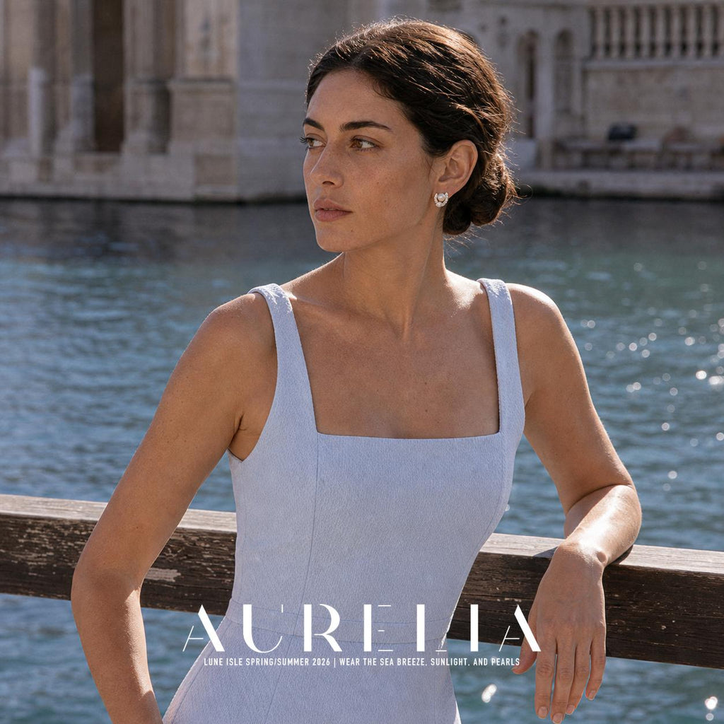 Model in a pale blue dress leaning against a waterside railing, wearing the same curved two-bead stud.