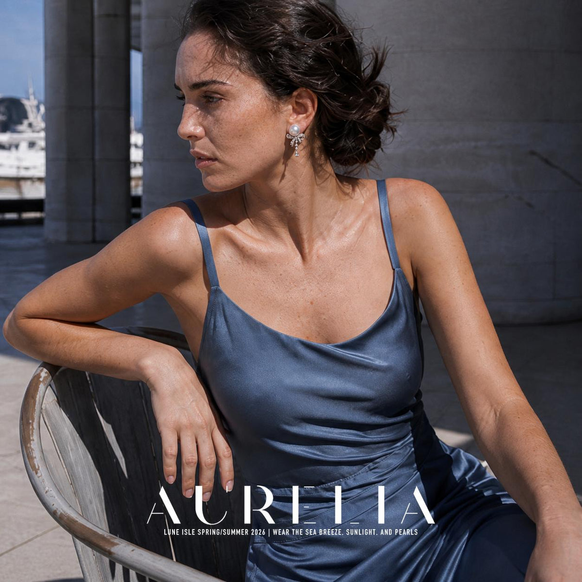 Seated person in a blue sleeveless dress wearing the pale round-top bow earring with a narrow clear-accent drop; outdoor columns are visible behind them.