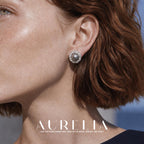Close view of a person wearing a round silver-colored stud with a dark gray round center and a clear faceted surround; reddish hair and a dark neckline are visible.