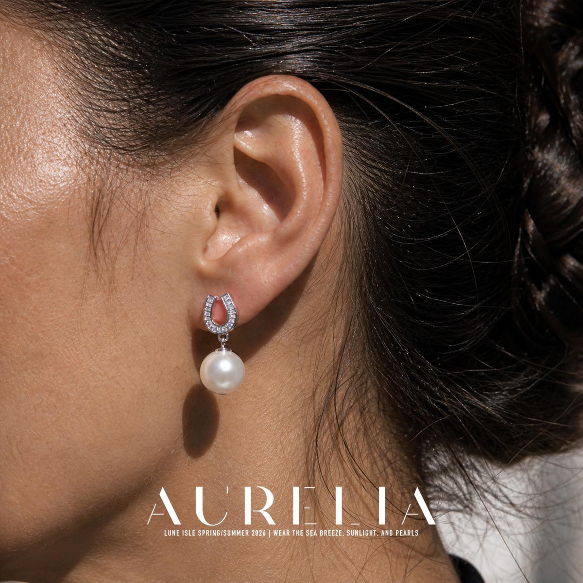 Close view of a person wearing a silver-colored earring with an open horseshoe-shaped top, narrow clear rectangular details and a white round drop.