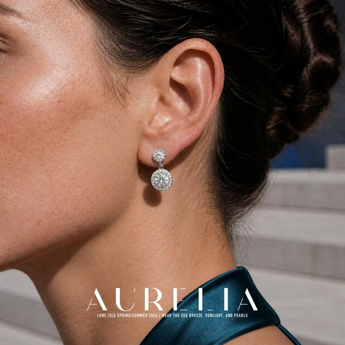 Close view of a person wearing a silver-colored earring with a clear round top linked to a larger round drop with a clear center and smaller clear accents around it; a dark teal collar is visible.