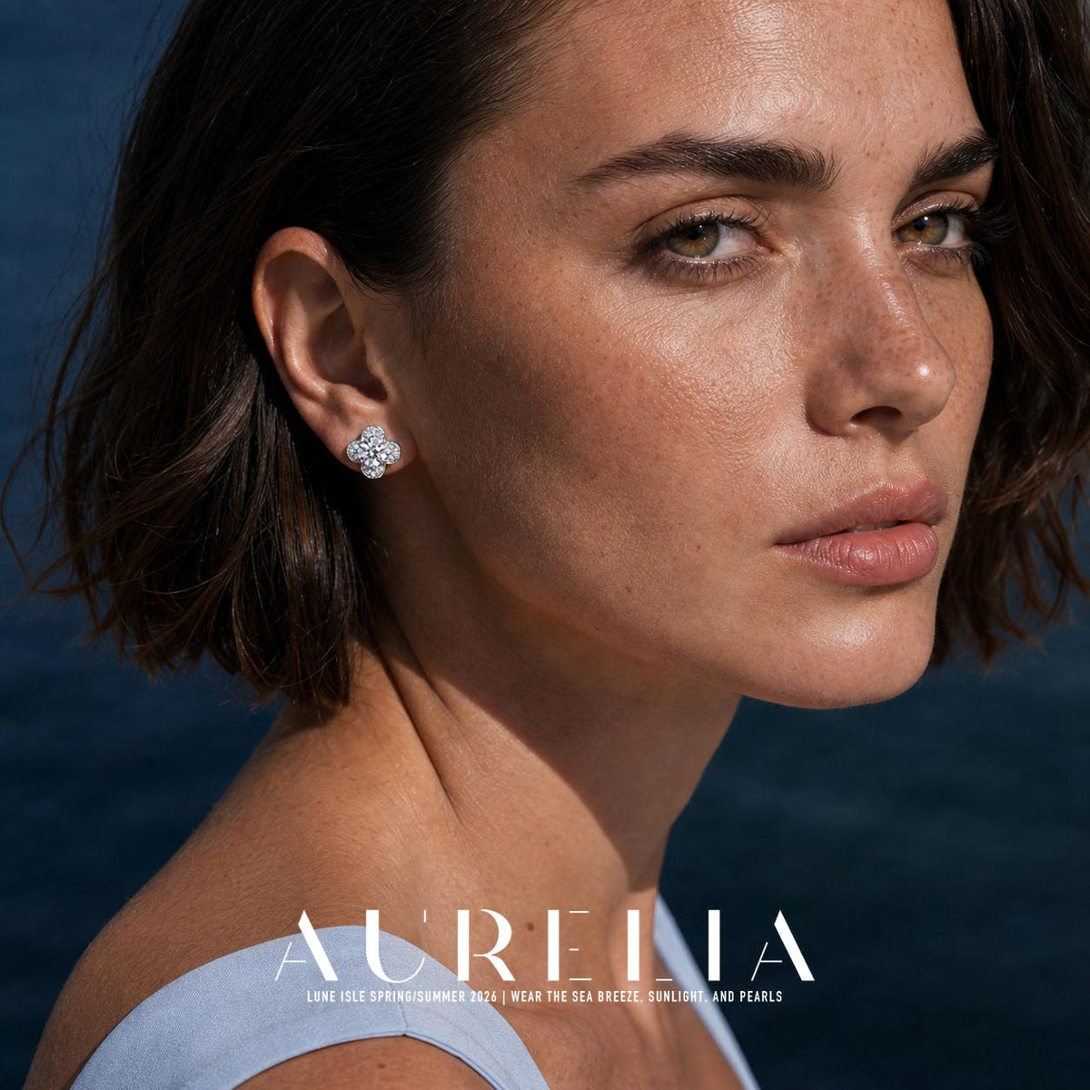 Over-shoulder portrait in a pale blue garment with the clover stud clearly visible.