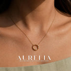 Close view of a gold-colored chain and open circular pendant worn at the upper chest, with one small clear round accent at the bottom inside the circle; a pale green neckline is visible.