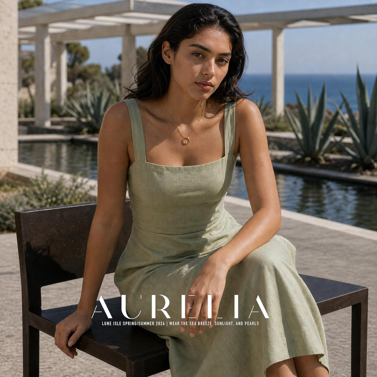 Seated person in a pale green sleeveless dress wearing a fine gold-colored chain and open circular pendant; a pool, plants and blue water are visible behind them.