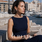 Seated person in a dark sleeveless dress holding a newspaper by a marina, wearing silver-colored hoops with paired white round lower elements.
