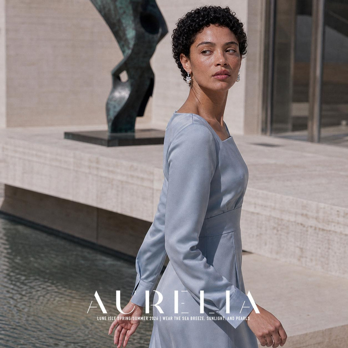 Person in a pale blue long-sleeved dress by a pool and abstract sculpture, wearing silver-colored bow-shaped drop earrings with pale round lower elements.