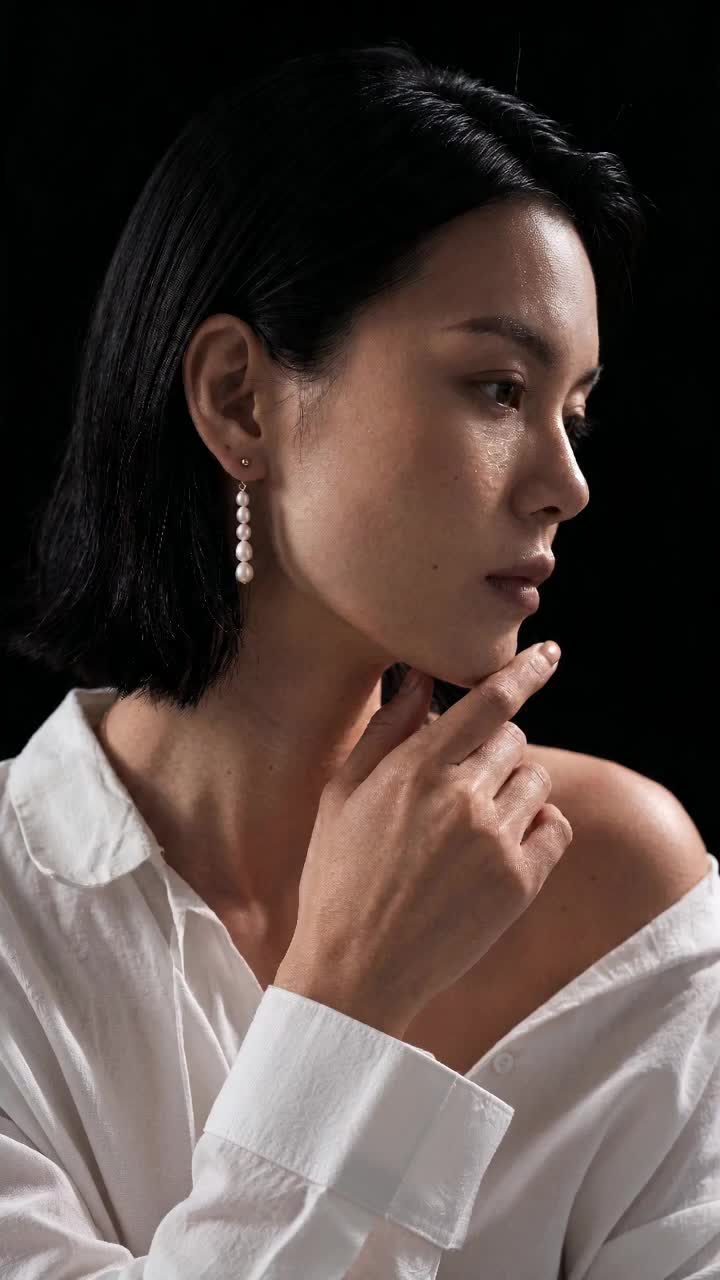 Narrow dark-background profile portrait in a white shirt; gold-tone ball stud and five graduated white beads visible, without visible branding.