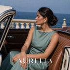 Model in a green dress seated in a car by the waterfront with the flower stud visible.